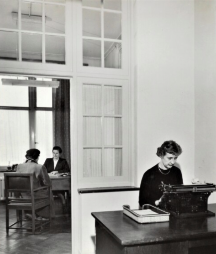 Op deze foto uit 1953 zien we het kantoor van het bureau van de sociale dienst van de Heerlense Vroedvrouwenschool ter ondersteuning van ongehuwde moeders. Een sociaal werkster is in gesprek met een cliënte. Op de voorgrond een typiste aan het werk. Harry Grubben fotoatelier - Fotocollectie SHCL.